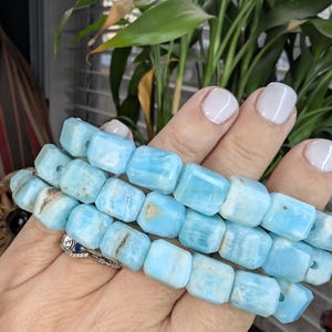 May include: Three strands of light blue gemstone beads. The beads are rectangular with rounded edges and have natural variations in color. The beads are held in a hand, with a ring visible on the finger.