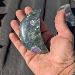 May include: A crescent-shaped stone with a dark green base color and purple and pink inclusions. The stone is held in a hand, showcasing its polished surface and natural variations. The overall shape resembles a moon.