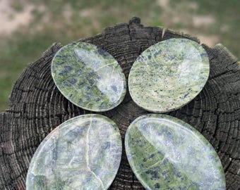 Puede incluir: Cuatro piedras verdes ovaladas con vetas verde oscuro y negro. Las piedras están dispuestas en un patrón circular sobre una superficie de madera marrón oscura y desgastada. El fondo es verde borroso.