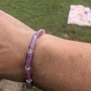 May include: A beaded bracelet featuring large, irregular, light purple beads. The bracelet is strung with clear, faceted beads between the larger beads. The bracelet is worn on a person's arm, with a blurred green background.