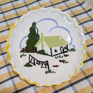 May include: A white ceramic pie plate with a scalloped edge, featuring a hand-painted cottage scene. The design includes a green tree, a house with a green roof, and a clothesline. The rim is trimmed with yellow paint.