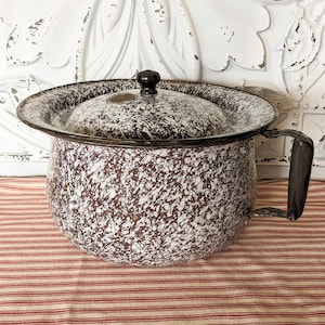 May include: A vintage enamelware chamber pot with a speckled brown and white pattern. It has a matching lid with a black knob and a dark handle. The pot sits on a red and white striped fabric.