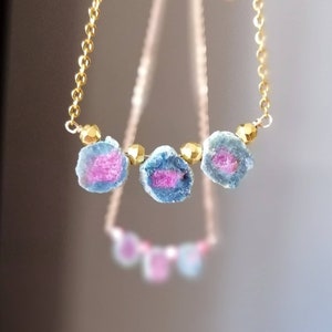 Puede incluir: Un collar de cadena dorada con tres colgantes de piedras preciosas rosas y azules. Las piedras preciosas tienen una forma irregular y una textura rugosa y natural.