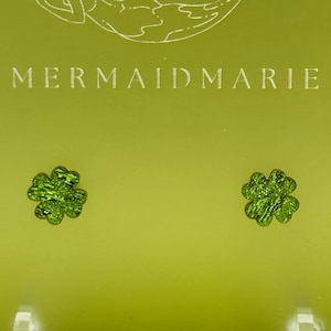 Puede incluir: Un par de pendientes de trébol de cuatro hojas verdes en un expositor con el texto "MERMAIDMARIE" en él.