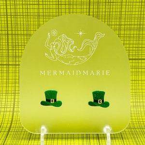 Puede incluir: Un par de pendientes de sombrero de duende verde con purpurina en un expositor verde claro con una ilustración de sirena y el texto "MERMAIDMARIE".