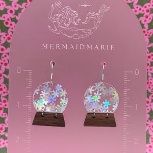 Op de afbeelding: Transparante, ronde oorbellen met iriserende sneeuwvlokaccenten en een donkere houten basis. De oorbellen hangen aan zilveren haken. De tekst "MERMAIDMARIE" is zichtbaar op de roze achtergrond.