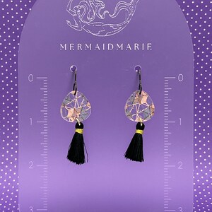 May include: Earrings with a geometric pattern in pink, blue, and gold, each with a black tassel and gold detail. The earrings are displayed on a purple stand. The text "MERMAIDMARIE" is visible on the stand.