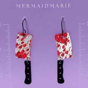 May include: A pair of novelty earrings shaped like cleavers. The blades are silver with red splatter, and the handles are black. The earrings hang from black hooks. The text "MERMAIDMARIE" is at the top.