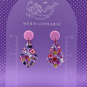 May include: A pair of dangle earrings with a floral and skeleton design. The earrings have a lavender background with colorful flowers and a skeleton. The earrings are displayed on a purple stand with the text "MERMAIDMARIE".