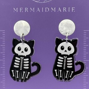 May include: A pair of black and white cat skeleton earrings. The earrings feature a black cat silhouette with a white skeleton design. The earrings have a white marbled top and are hanging on a purple background. The text "MERMAIDMARIE" is at the top.