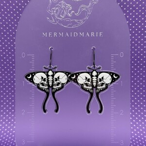 May include: A pair of black and white moth-shaped earrings with skull designs. The earrings are displayed on a purple stand with the text "MERMAIDMARIE". The earrings have a unique, gothic aesthetic.