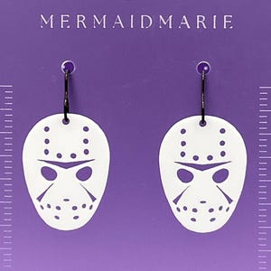May include: White acrylic earrings featuring a hockey mask design with black ear wires. The earrings are in the shape of a hockey mask with eye and mouth cutouts and dot details. The earrings are on a purple background with the text "MERMAIDMARIE".