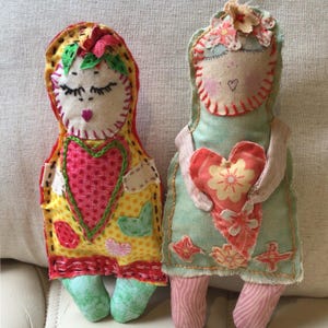 May include: Two handmade fabric dolls. One doll wears a yellow dress with a pink heart and green feet. The other doll wears a mint green dress and holds a heart with a floral design. Both dolls have closed eyes and floral headbands.