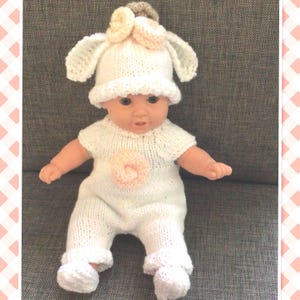 Könnte beinhalten: Eine Puppe in einem handgestrickten weißen Outfit, bestehend aus einem langärmeligen Strampler mit einer blassrosa Blumenapplikation auf der Brust, einer passenden weißen Mütze mit zwei großen Schlappohren und einer blassrosa Blume sowie weißen Strickschuhen.