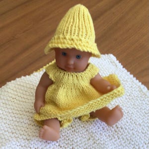 May include: A small doll wearing a handmade yellow knitted dress and matching hat. The doll has blue eyes and is sitting on a white knitted blanket. The dress has a ruffled hem and cap sleeves.