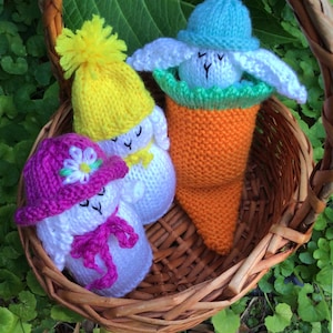 Puede incluir: Una cesta de mimbre contiene tres figuras de conejitos de Pascua de punto. Uno está dentro de una zanahoria naranja, otro lleva un sombrero amarillo con un pompón y el tercero tiene un sombrero morado con una flor. Todos son blancos con ojos negros.