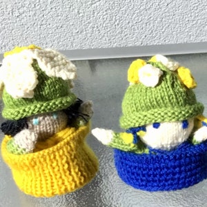 May include: Two knitted dolls with green hats and white flower accents. One doll is wearing a yellow knitted dress and the other is wearing a blue knitted dress.