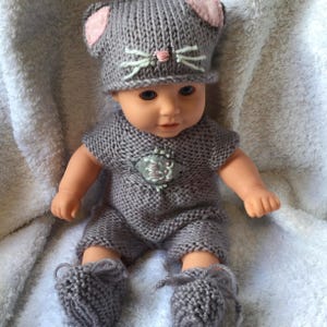 May include: A doll wearing a gray knitted outfit, including a hat with pink ears and embroidered whiskers, a short-sleeved romper, and booties. The outfit features a decorative patch on the chest. The doll is sitting on a white, textured surface.