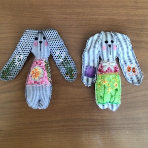 May include: Two handmade fabric bunny dolls. One bunny has blue and white polka dot ears and a striped body, with a pink floral dress. The other bunny has striped ears and body, with a green floral dress. Both have embroidered details.