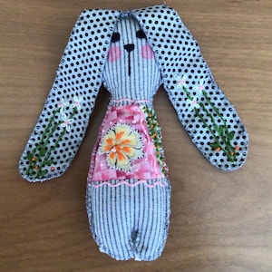 May include: A fabric rabbit doll featuring long ears with a black and white polka dot pattern and green embroidered floral stems. The body is made of grey striped fabric with a pink checkered patch on the chest, a yellow flower applique, and pink cheeks.