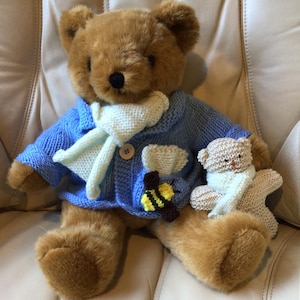 Puede incluir: Un oso de peluche marrón con un cárdigan de punto azul claro y una bufanda blanca, con un pequeño motivo de abeja. Un oso de peluche más pequeño, tejido, con una bufanda blanca está a su lado. Los osos están sobre una superficie crema.