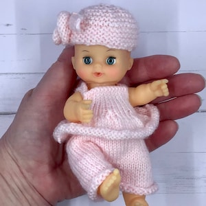 May include: A small pink knitted doll with blue eyes and a pink knitted hat. The doll is wearing a pink knitted dress and pants.