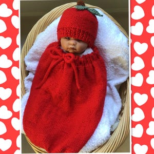 May include: A red knitted baby outfit, resembling a tomato, includes a hat with a green leaf and a matching sack. The baby is nestled in a wicker basket lined with a white towel. The outfit is handmade.