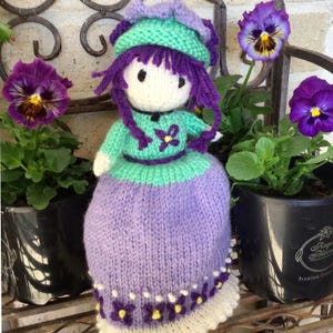 May include: A knitted doll with purple hair, a green dress, and a purple hat. The doll is sitting on a shelf with two potted plants with purple flowers.