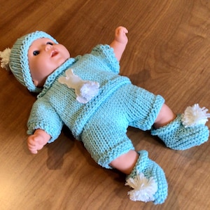 May include: A baby doll wearing a light blue knitted outfit, including a hat, sweater, shorts, and booties. The sweater features a white bunny applique. Each piece has white pom-pom accents. The doll is lying on a wooden surface.