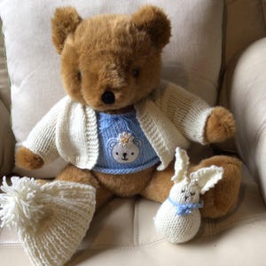 May include: A brown teddy bear wearing a blue sweater with a bear face and a white cardigan. A white knitted hat with a pom-pom and a small white knitted bunny with a blue ribbon are also present.