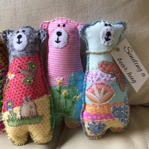 May include: Three handmade fabric bear dolls in red, pink, and blue. Each bear has a white face with a black nose and stitched details. The bears have patchwork bodies with floral and butterfly designs. One bear has a tag that says "Sending a bear hug."