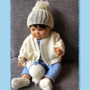 May include: A knitted baby doll wearing a white knitted hat with a white pom-pom, a white knitted cardigan, blue knitted pants, and white knitted booties. The doll is holding a white knitted toy.