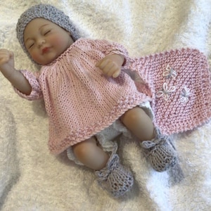 May include: A baby doll dressed in a light pink knitted dress, matching hat, and grey booties. A matching pink blanket with white embroidered flowers is next to the doll. The doll is lying on a white, textured surface.