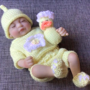 May include: A yellow knitted baby doll outfit with a matching hat and a yellow knitted duck toy. The outfit features a flower design on the front.