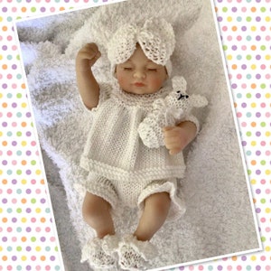 May include: A white knitted baby doll outfit with a matching hat and booties. The outfit includes a dress with a ruffled hem, shorts, and a bow on the hat. The doll is holding a white knitted bunny.