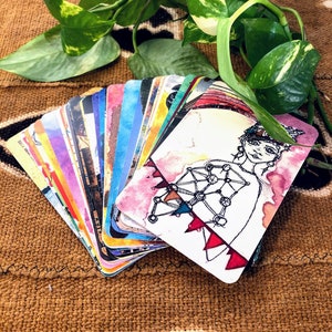 May include: A fan of rectangular cards featuring colorful watercolor illustrations and black ink line drawings. The top card displays a figure with a geometric chest design and red triangular accents, while the other cards show various abstract patterns.