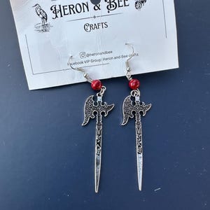 Può includere: Orecchini color argento con un design a spada e ali, ciascuno con una piccola perla rossa. Gli orecchini sono esposti su una carta bianca con la scritta "HERON & BEE CRAFTS".