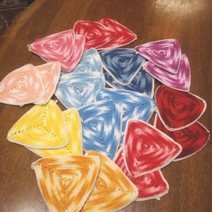 Può includere: Una collezione di presine triangolari fatte a mano, all'uncinetto, in vari colori, tra cui rosso, rosa, blu, giallo e arancione. Ogni presina presenta un design a spirale e un bordo bianco, ideale per la cucina.