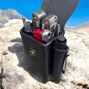 Puede incluir: Una funda de cuero negro para multiherramientas con varias herramientas plateadas y rojas en su interior. La funda tiene un cierre de botón a presión y una solapa de cuero negro. Una pequeña linterna negra está sujeta al lateral. El fondo muestra una cordillera y cielo azul.