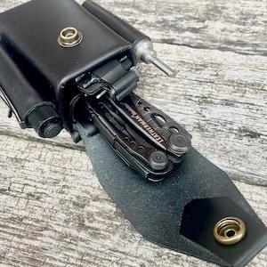 Puede incluir: Funda negra de cuero para multiherramientas con una multiherramienta Leatherman parcialmente visible. La funda tiene un cierre a presión y un pequeño bolsillo para un bolígrafo u otra herramienta. La multiherramienta es plateada y negra.