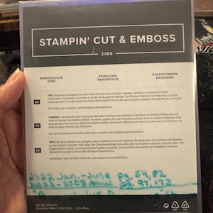 May include: A Stampin' Cut & Emboss die set package. The package is black and white with the words "STAMPIN' CUT & EMBOSS" at the top. The package includes the words "RANUNCULUS DIES" and "POINÇONS RENONCULE". The package dimensions are 5.67 x 4.88 inches.
