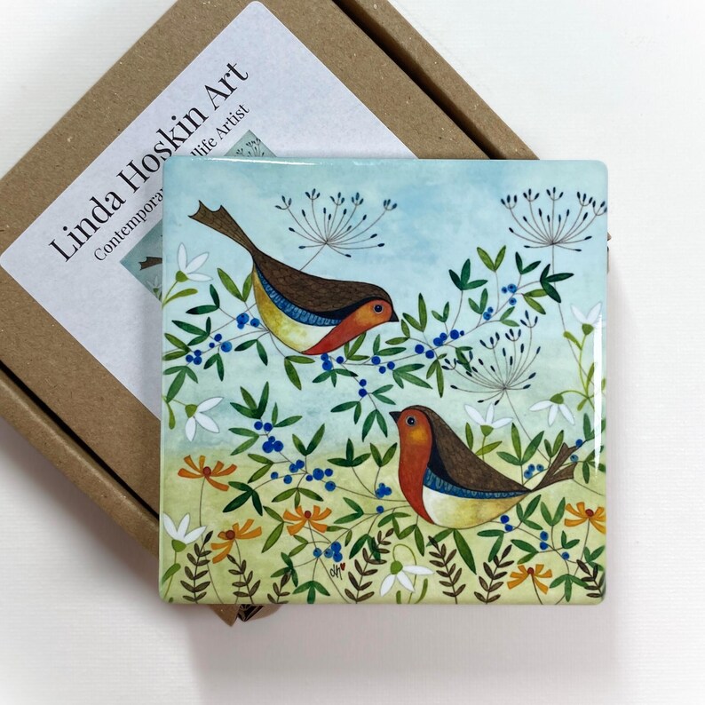May include: A square ceramic tile featuring a watercolour painting of two robins perched on a branch with blue berries. The background is a light blue sky with white flowers and green leaves.