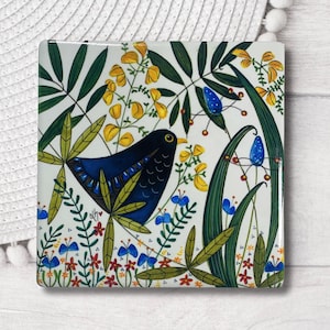 May include: A square ceramic tile featuring a hand-painted illustration of a dark blue bird perched among vibrant green foliage, yellow blossoms, and small blue flowers. The artwork includes fine line details and a white background.