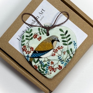 May include: A white ceramic heart-shaped ornament with a hand-painted illustration of a bird with blue, yellow, and white feathers surrounded by red berries and green leaves. The ornament has a brown twine loop for hanging. The text "Linda Hoskin Art Wildlife Artist" is printed on the box.