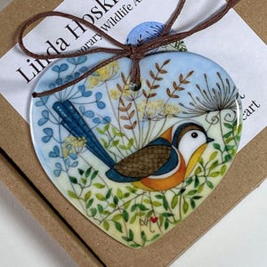 May include: Heart-shaped ceramic ornament with a bird illustration. The bird has a white face, brown back, and orange chest, set against a blue sky and green foliage. A brown cord is attached for hanging. The ornament is approximately 10 cm in width.