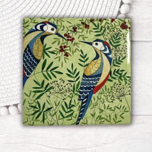 May include: Square ceramic coaster with a colourful illustration of two woodpeckers amongst green foliage and red berries. The birds have blue, yellow, and red markings. The background is a light green colour.