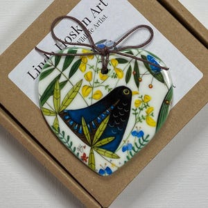 May include: A white ceramic heart-shaped ornament featuring a blue and black bird, green leaves, and yellow flowers. The ornament has a hole at the top for hanging and says "Linda Hoskin Art Wildlife Artist" on the back.
