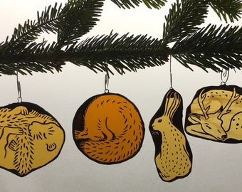 Puede incluir: Cuatro adornos navideños hechos a mano con diseños de animales. Los adornos son amarillos con contornos negros y representan un erizo, un zorro, un conejo y un ciervo. Cuelgan de una rama de pino verde con ganchos plateados.