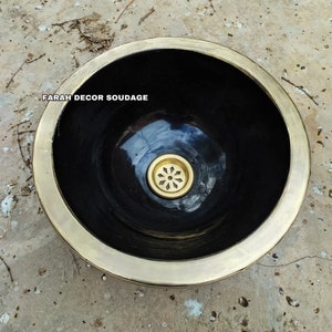 Puede incluir: Un lavabo de cerámica negra con un borde dorado y un desagüe dorado. El lavabo está sobre una superficie de hormigón. "FARAH DECOR SOUDAGE" está escrito en el lavabo.