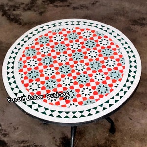 May include: A round, white table with a black metal base. The table top is decorated with a geometric pattern of red, green, and white tiles. The text "Farah decor Soudage" is visible on the table top.